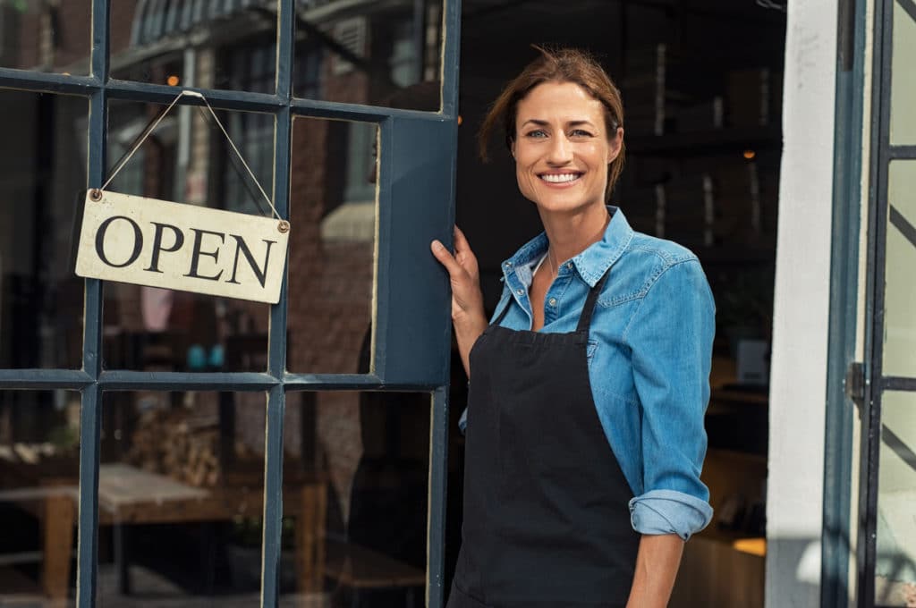 Portrait of a happy waitress standing at restaurant entrance. Portrait of mature business woman in black apron ready to attend new customers in her just opened coffee shop. Happy beautiful woman owner showing open sign in her small business shop.